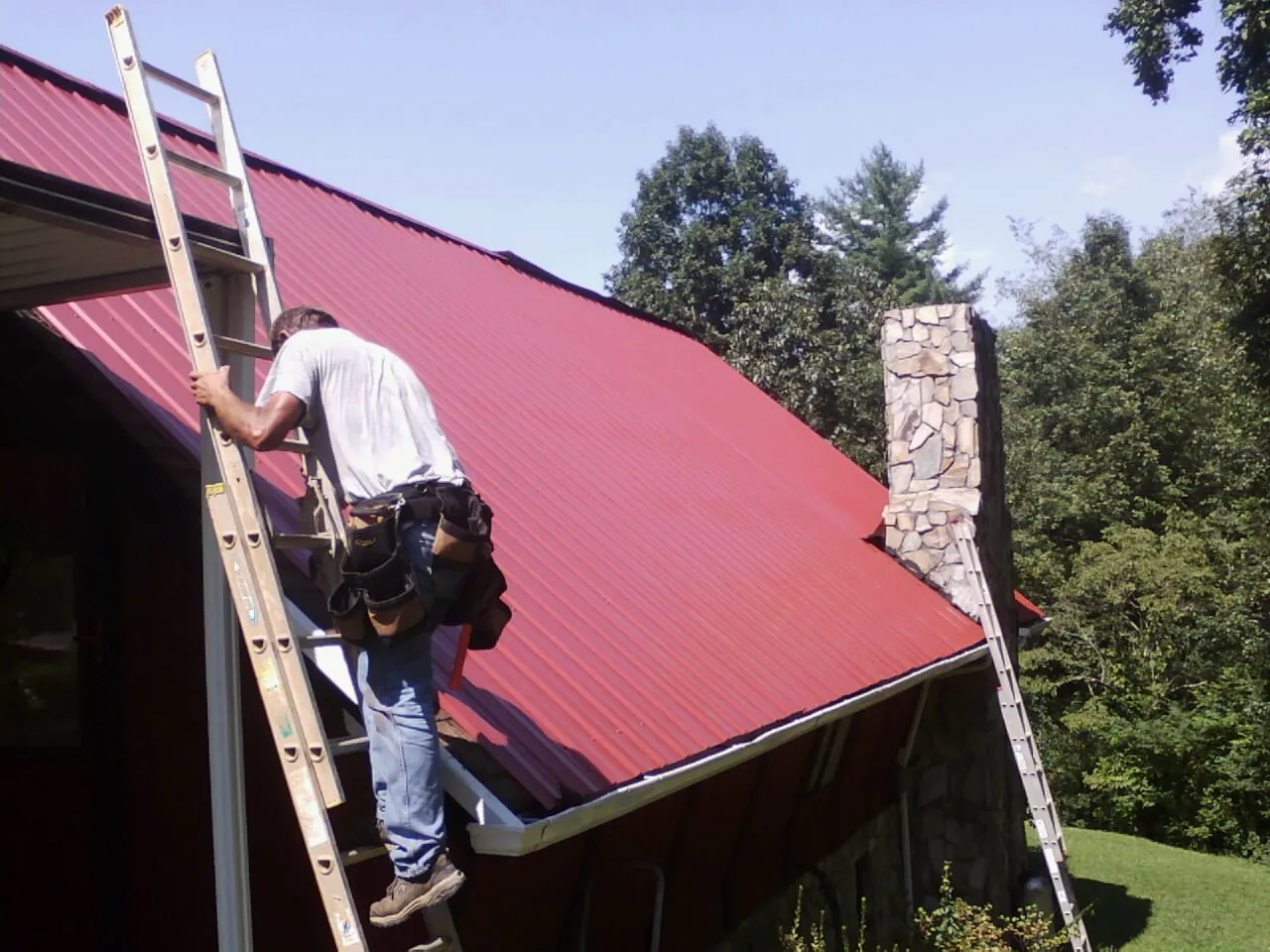 Roofer on a ladder beside a red metal roof and stone chimney — Soffit & Fascia Repair by Roofing Brothers in Franklin Park